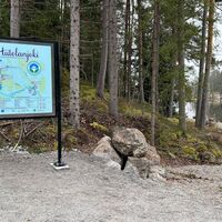 Informationsschild des Erholungsgebiets vor Bäumen, rechts ein Wegweiser, von dem ein Pfad zum Flussufer führt, der Fluss ist im Hintergrund zu sehen