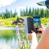 Eine junge Frau mit Hut sieht auf ihr Handy, in dem man den Hebalmsee sehen kann.