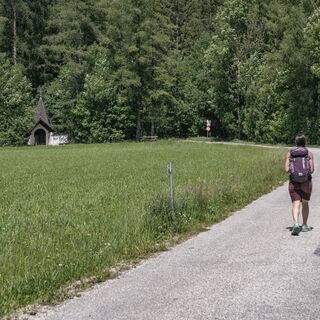 Das Bild zeigt eine Frau, die auf einer Asphaltstraße geht. Links von ihr ist eine Wiese, auf der eine Kapelle steht. Der Weg endet in einem Wald.