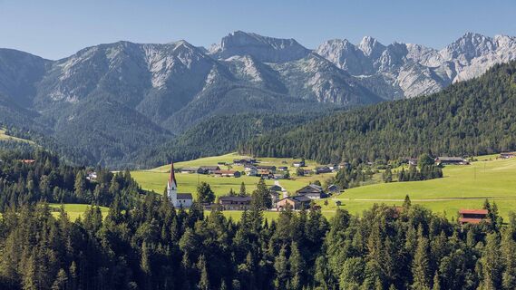 Blick auf das Dorf Steinberg am Rofan mit Kirche vor bewaldeten Hängen und felsigem Gebirge.